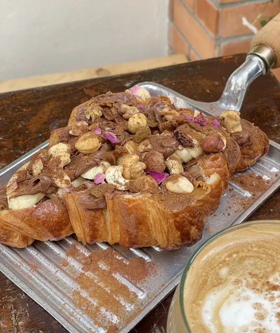 Un croissant garni de pâte à tartiner, fruits secs et pétales de rose, avec un cappuccino